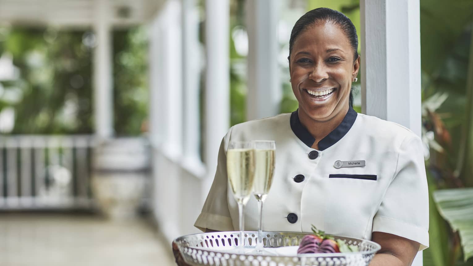Smiling hotel staff holds tray with two glasses of Champagne on veranda