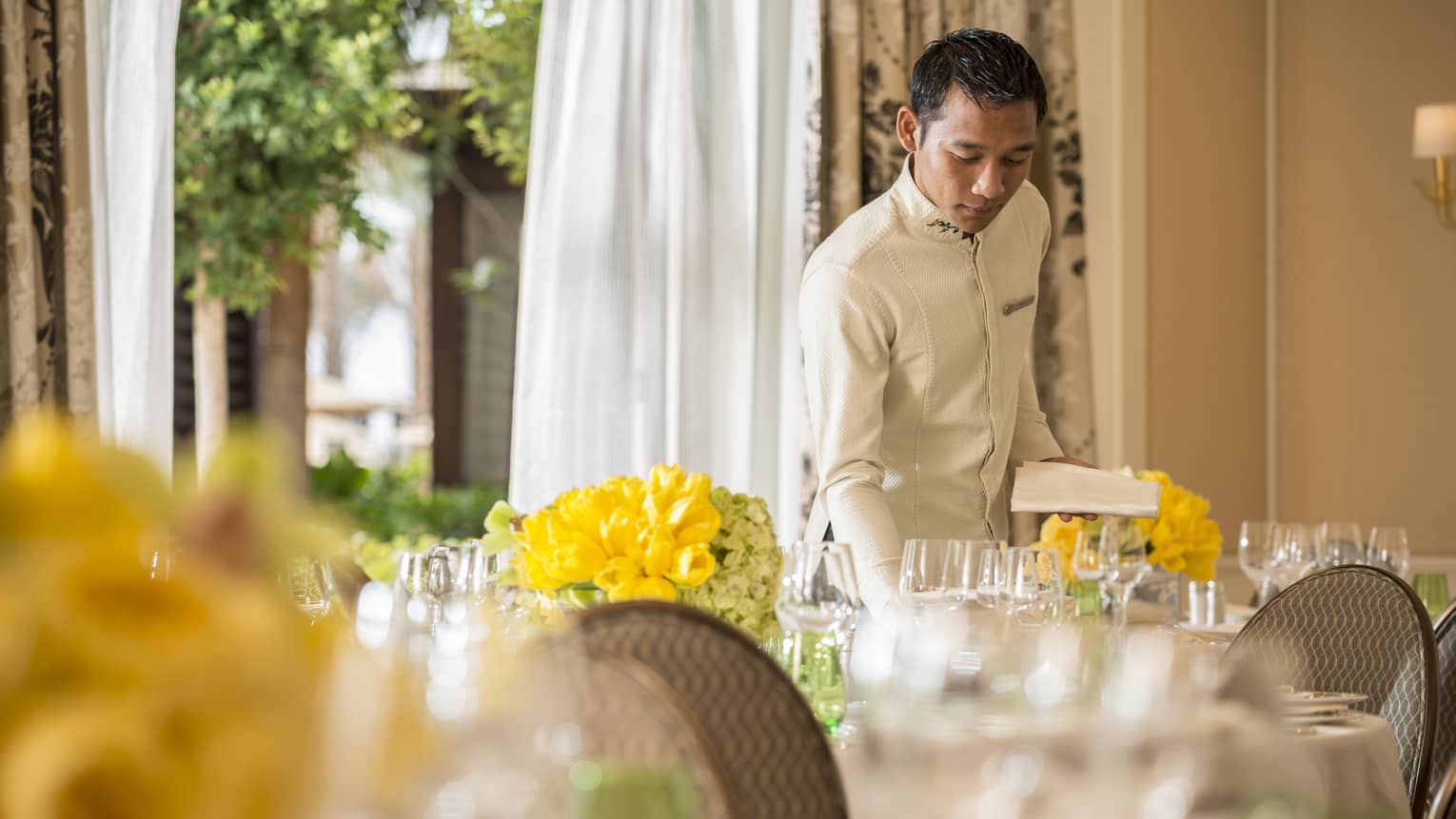 Hotel staff sets banquet table with wine glasses, yellow flowers in sunny event space 