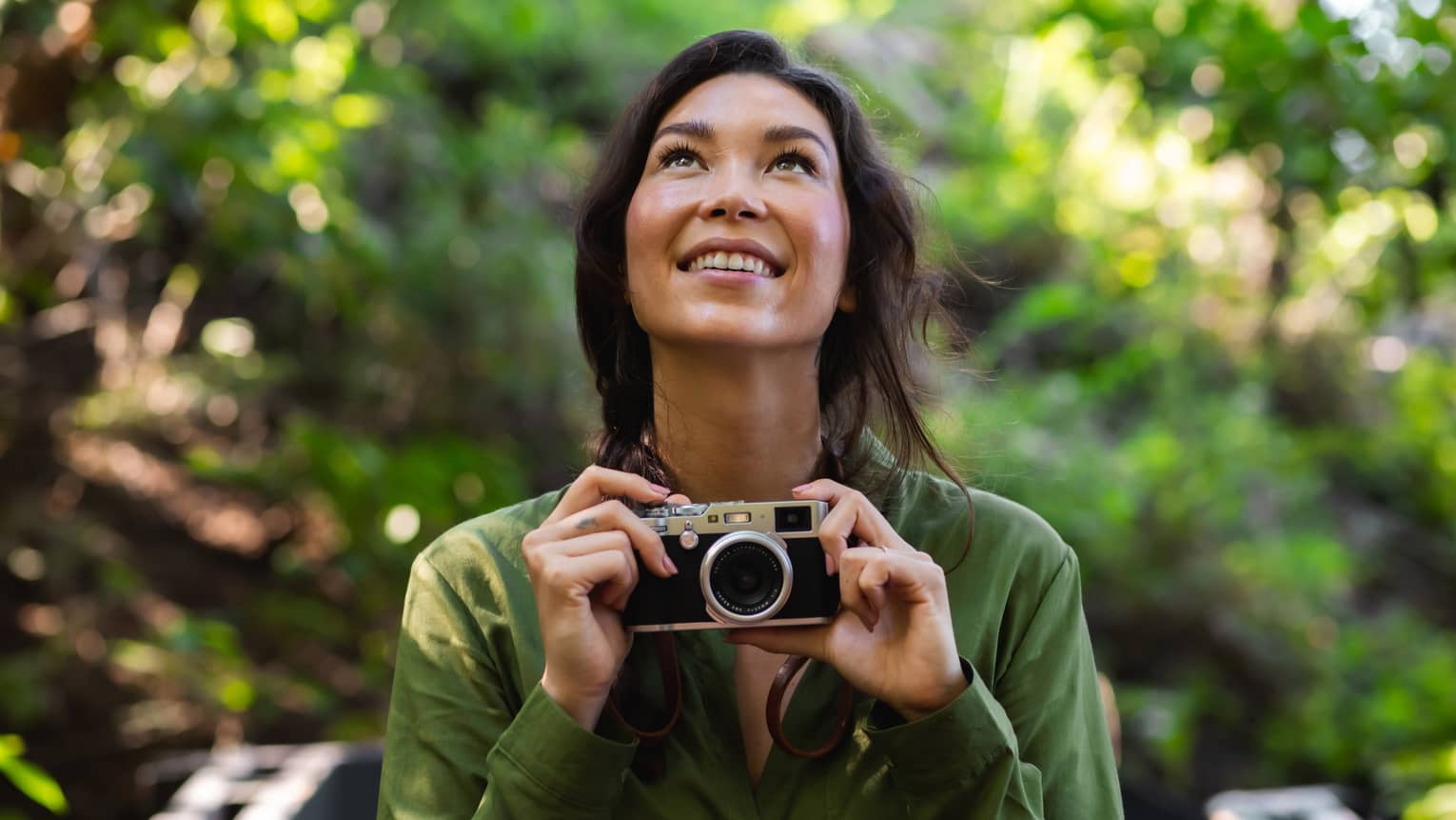 Woman with long dark hair wearing a green button-down shirt holds a camera as she looks upward in the forest