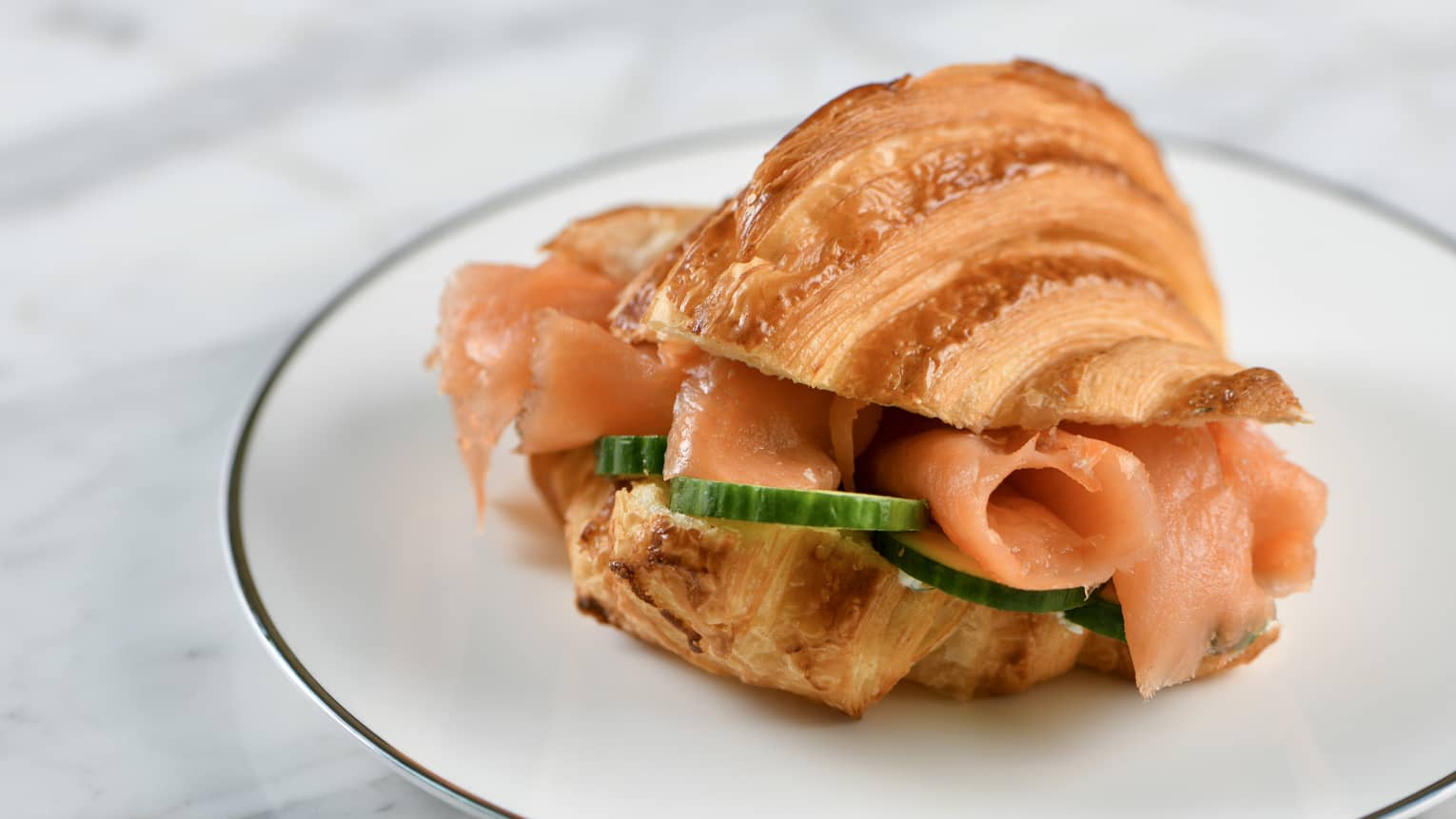 Salmon and cucumber slices in a croissant placed on a white, silver-lined plate