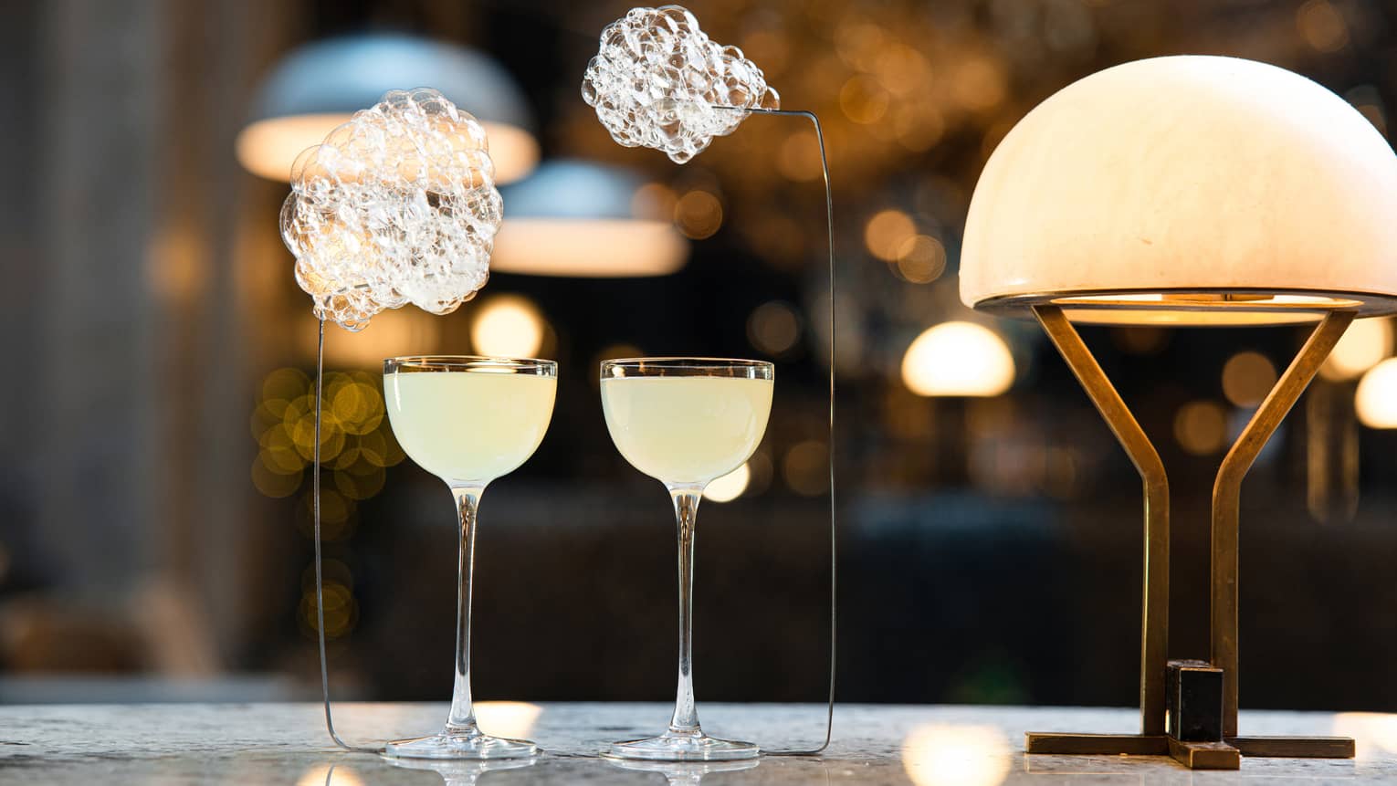 Two cloudy cocktails in unique glasses beside lamp on bar