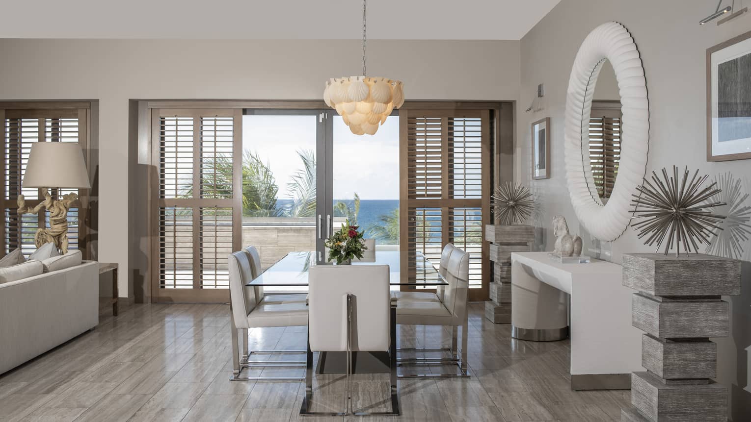Luxury resort penthouse dining area with a glass table, white leather chairs, coastal decor and large windows with wooden shutters offering an ocean view