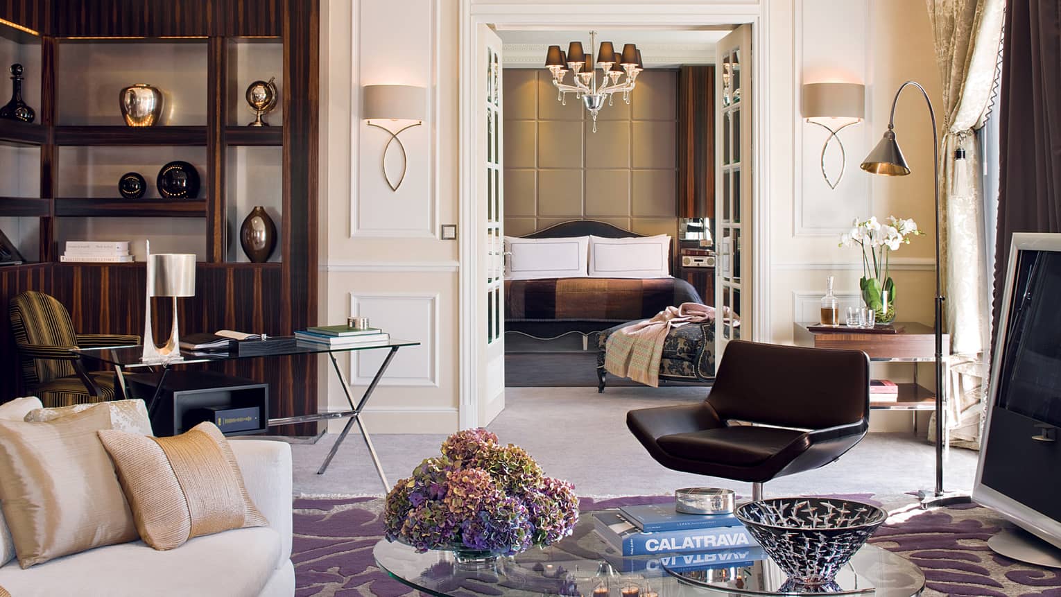 Presidential suite living area with sleek dark furniture, a glass coffee table, beige sofa, desk and decorative shelves, leading into a luxury bedroom