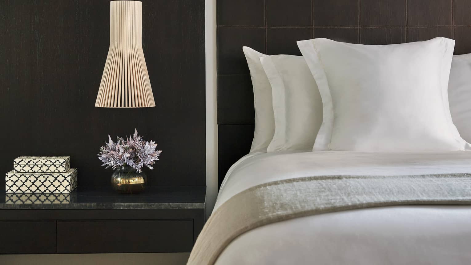 Close up of a nightstand with two small decorative boxes, small vase of flowers and a lamp next to a bed made with all-white linens and a dark wood headboard