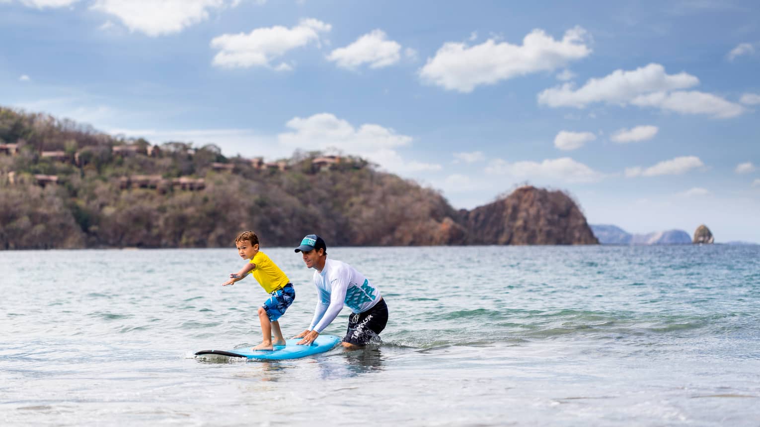 Man wading in ocean helps young child stand, balance on surfboard, mountains in background