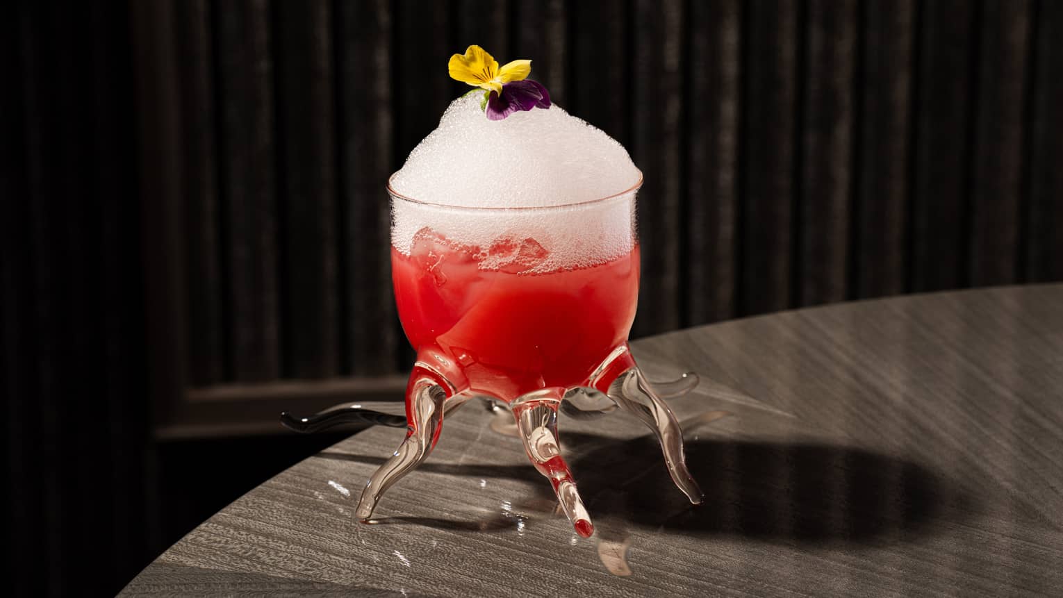 Red-hued cocktail topped with white foam and a yellow and purple flower served in a glass with multiple tentacle-shaped stems