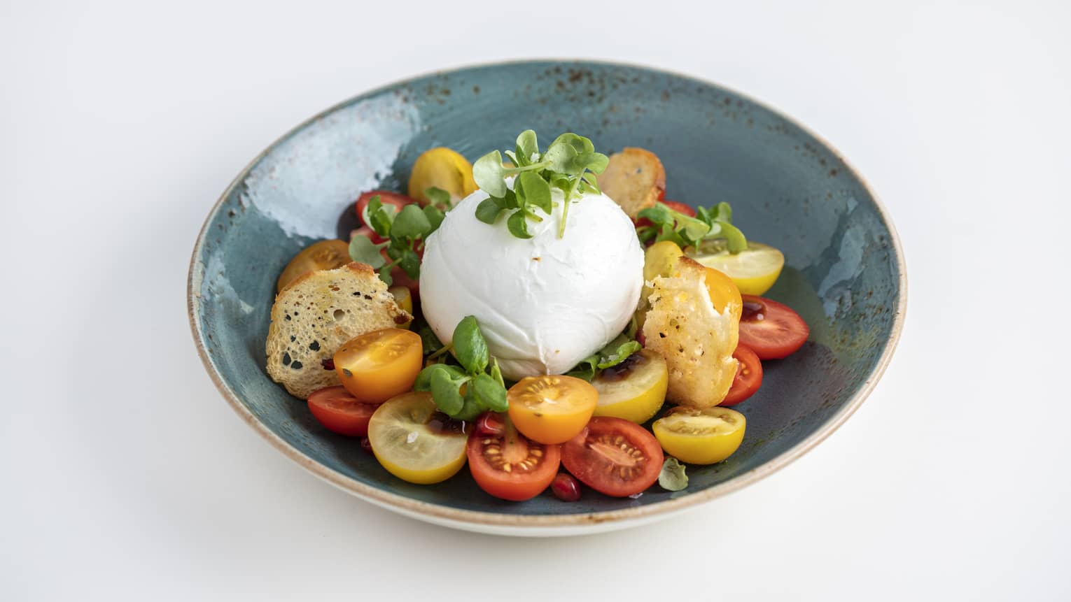 Burrata served with cherry tomatoes, arugula, pomegranate, balsamic drizzle and basil pesto in a round blue bowl