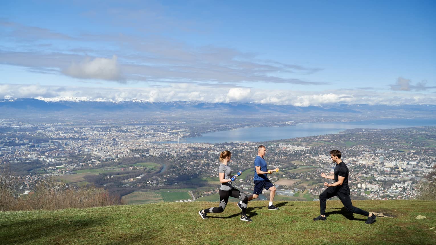 Two people on green mountainside doing knee bends with trainer and blue skies in background
