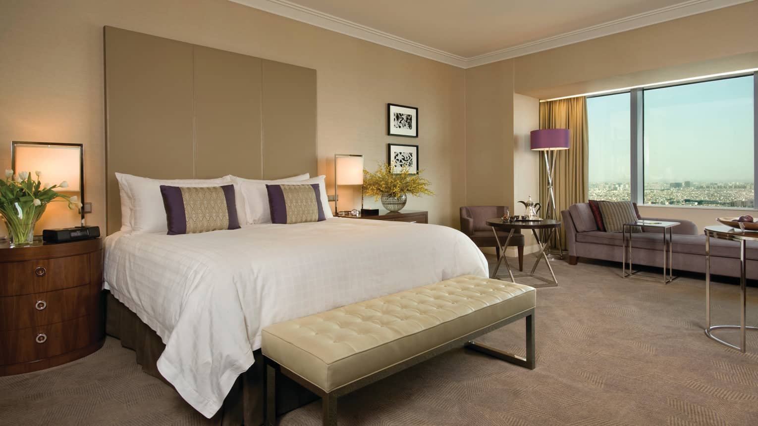 Deluxe hotel room bed with tall padded headboard and leather bench, beige carpet, long chaise at window