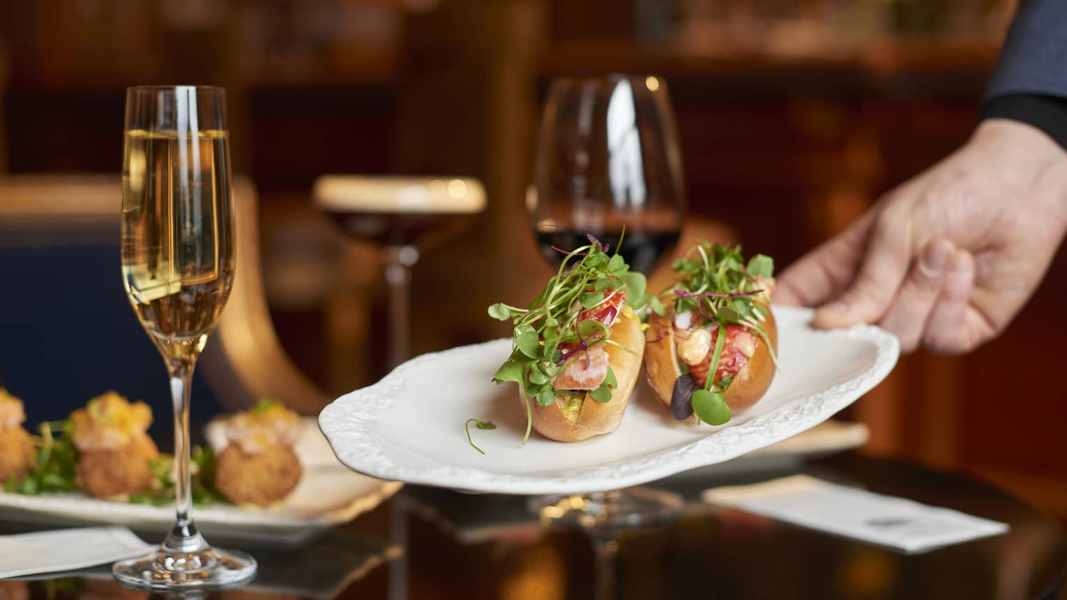 Two lobster rolls on a white plate being set on a dining table