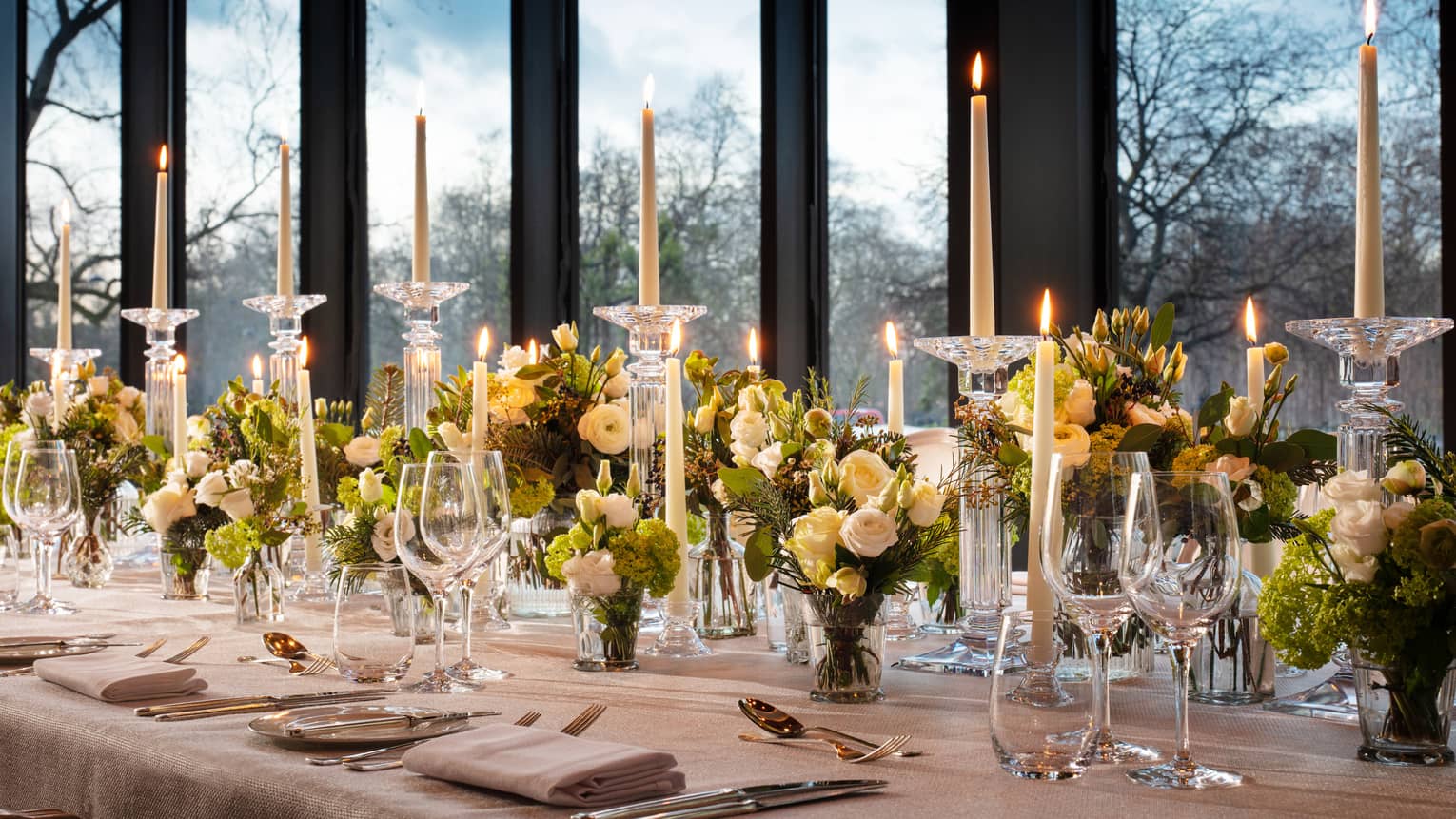 Closeup shot of meeting room table set with tall tapered candles and green floral arrangements against a window backdrop