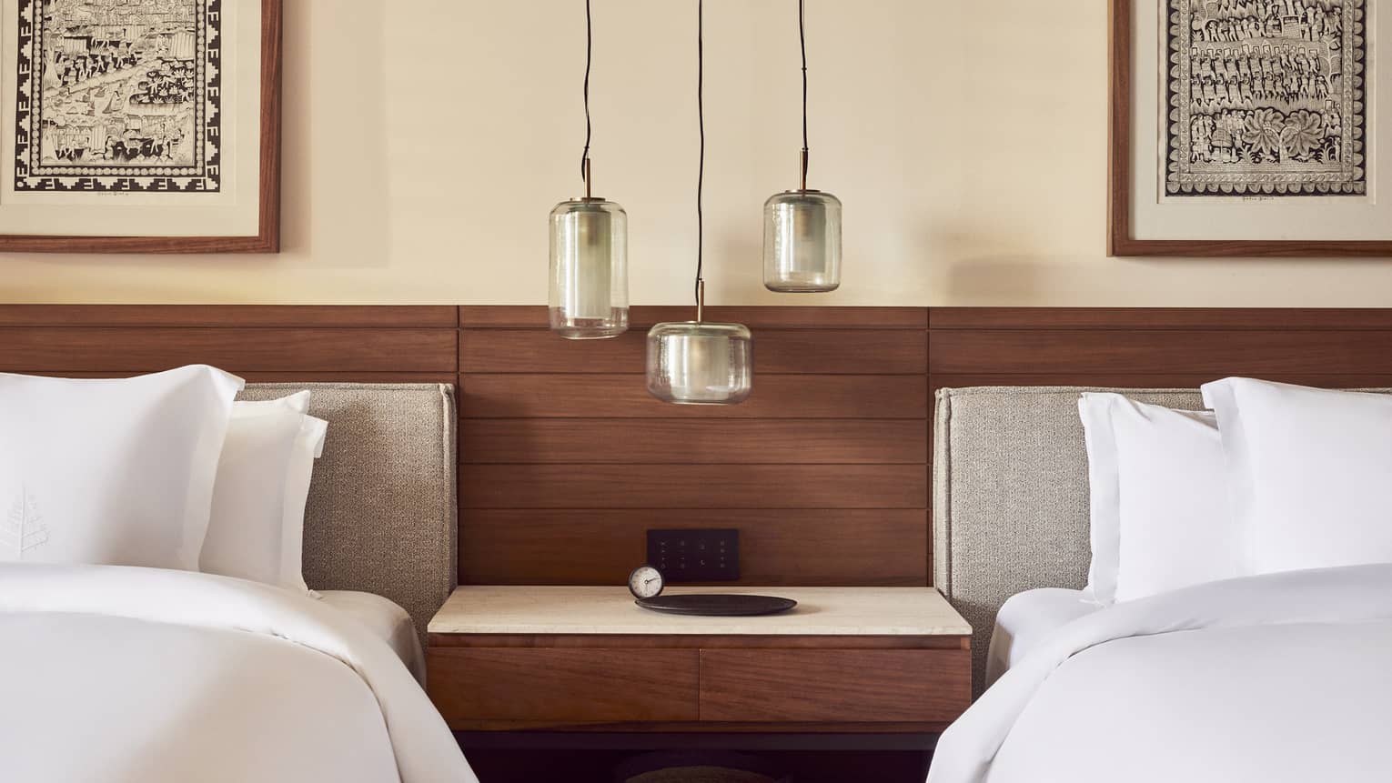 Cozy bedroom with two beds featuring white linens, a wooden headboard and a shared nightstand. Three pendant lights hang above, framed by artwork on the walls.
