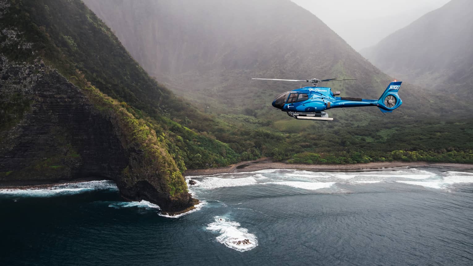 A bright blue helicopter flying above dark blue ocean waters with a verdant, mountainous coastline and misty skies in the background
