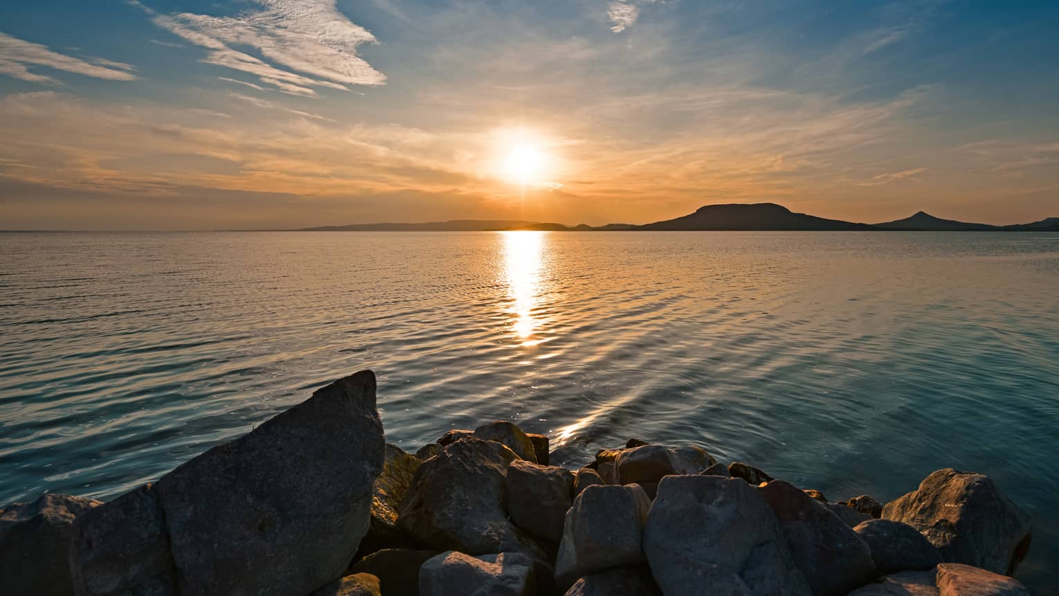 A jagged rocky shoreline looking out across the lake at hills being silhouetted by the setting sun.