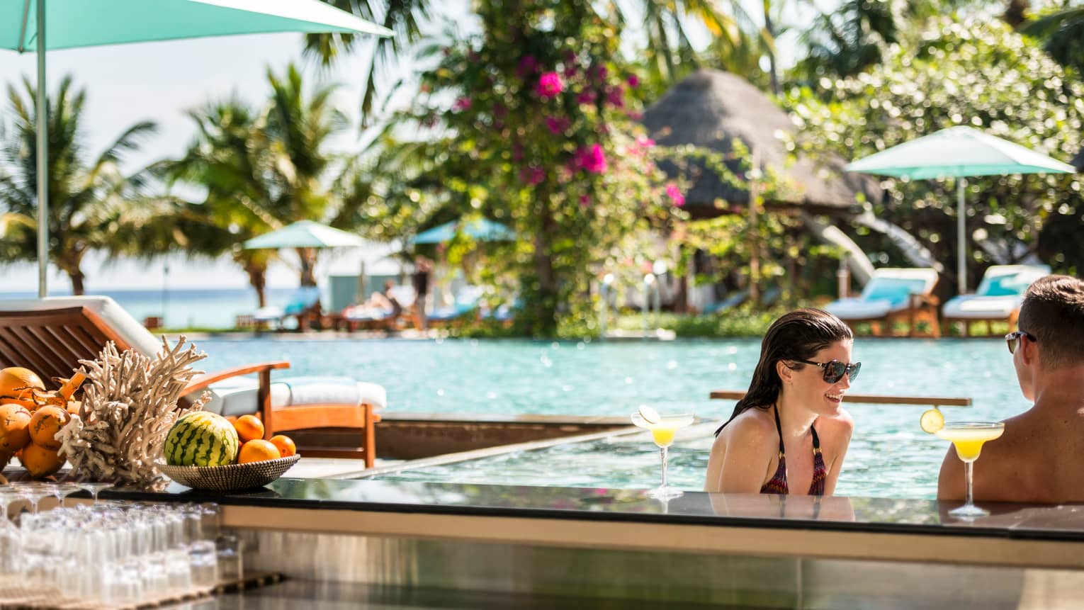 Woman, man wade in outdoor swimming pool with tropical cocktails, baskets of fresh fruit on ledge