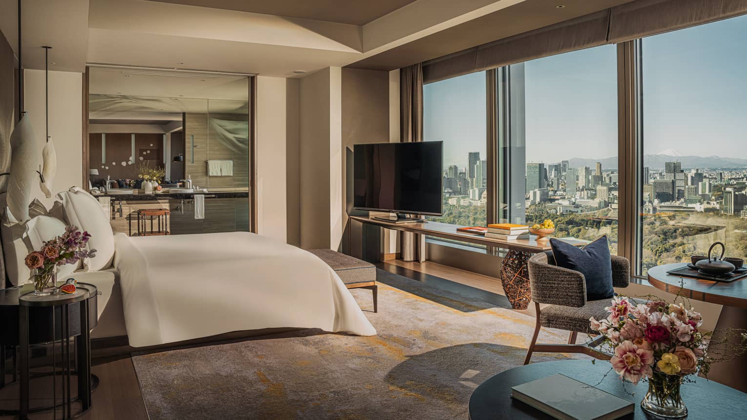 Luxury hotel suite featuring a king bed facing a wall of windows, plus a dining table