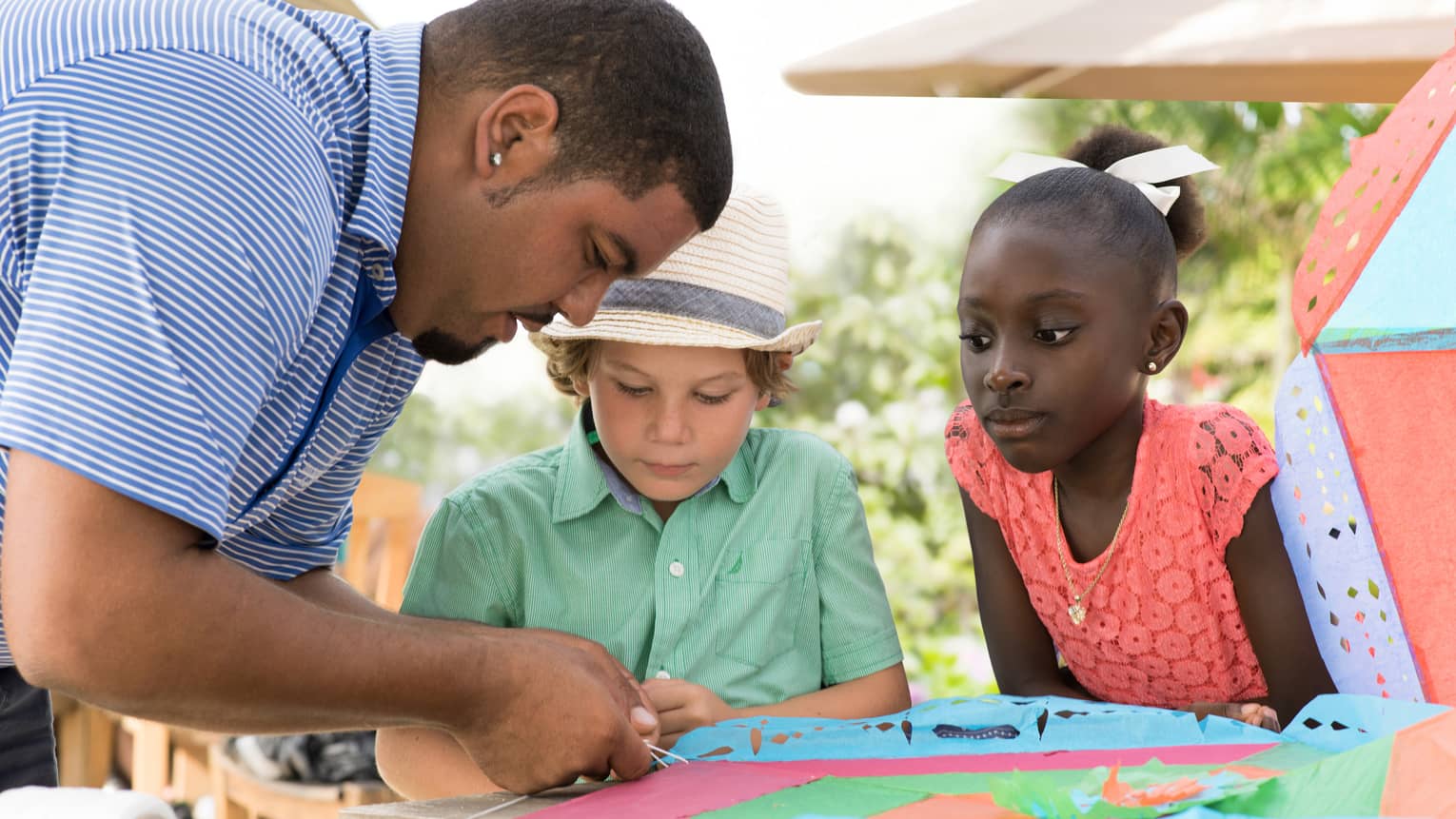 Man ties string onto colourful paper kite at table as two children watch