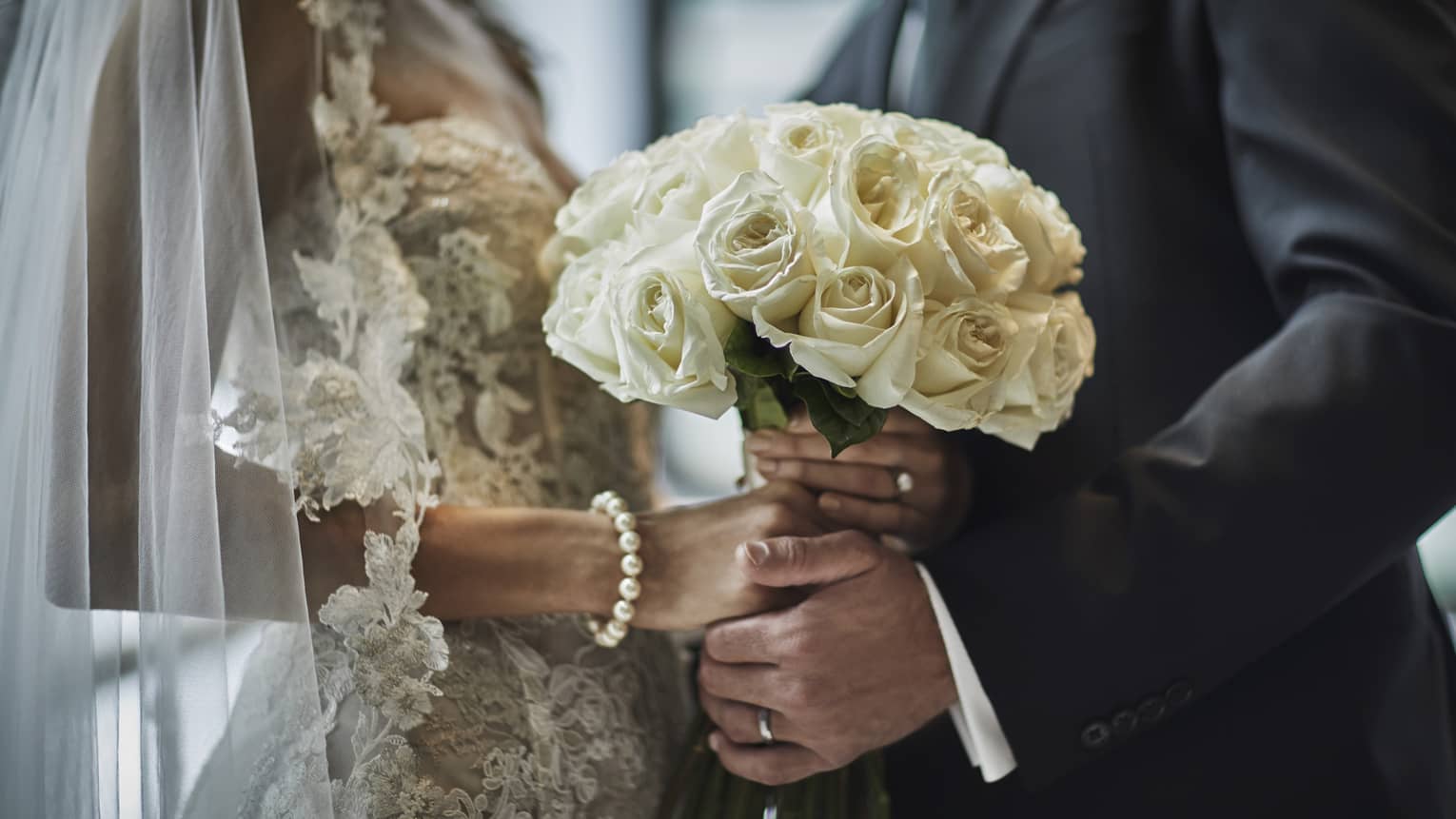 A close up of a bride in a lace gown and a groom in a gray suit hold the bride's bouquet of white roses together.