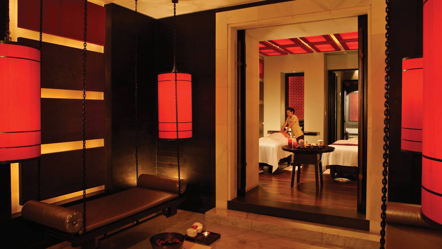 Spa staff gives massage on table behind doorway in dark spa with hanging red lanterns