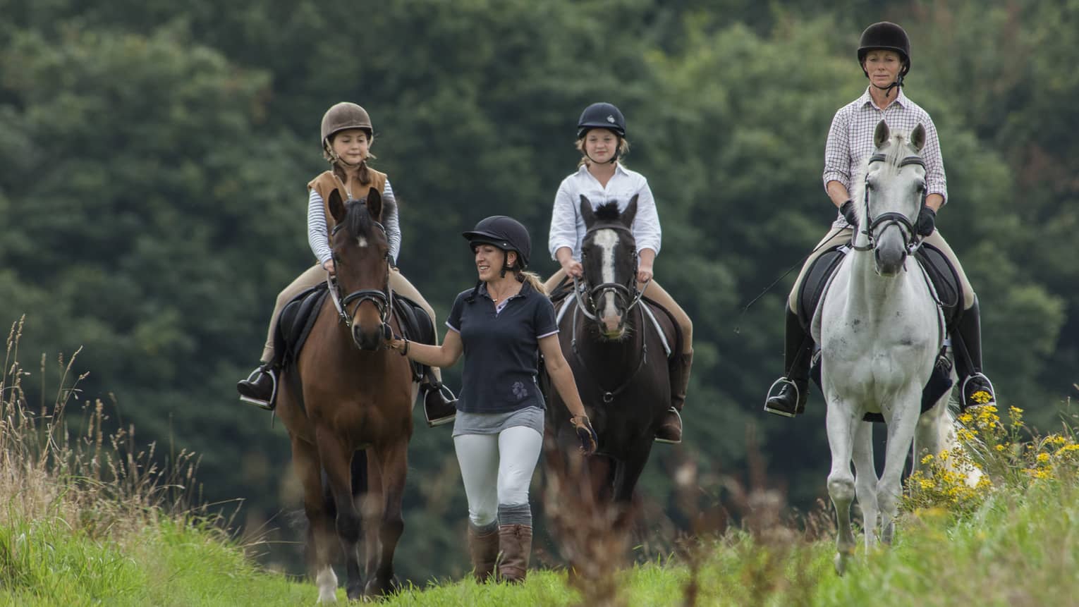 Three children wearing equestrian-style helmets, riding on horseback and led by trainer