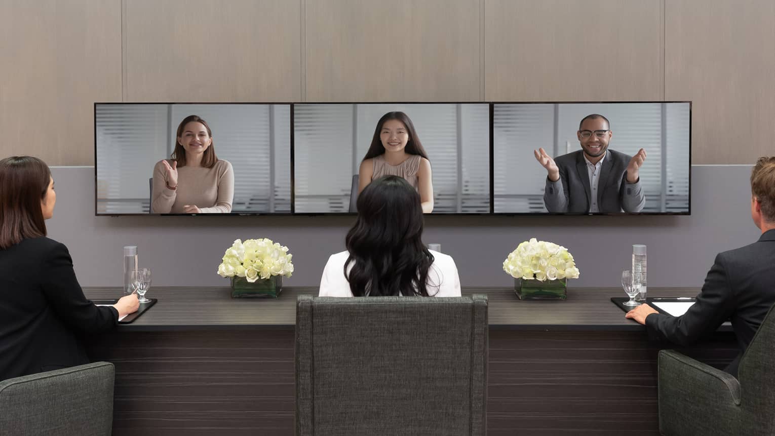 Several people in business clothes sit in front of a series of screens in a virtual meeting.