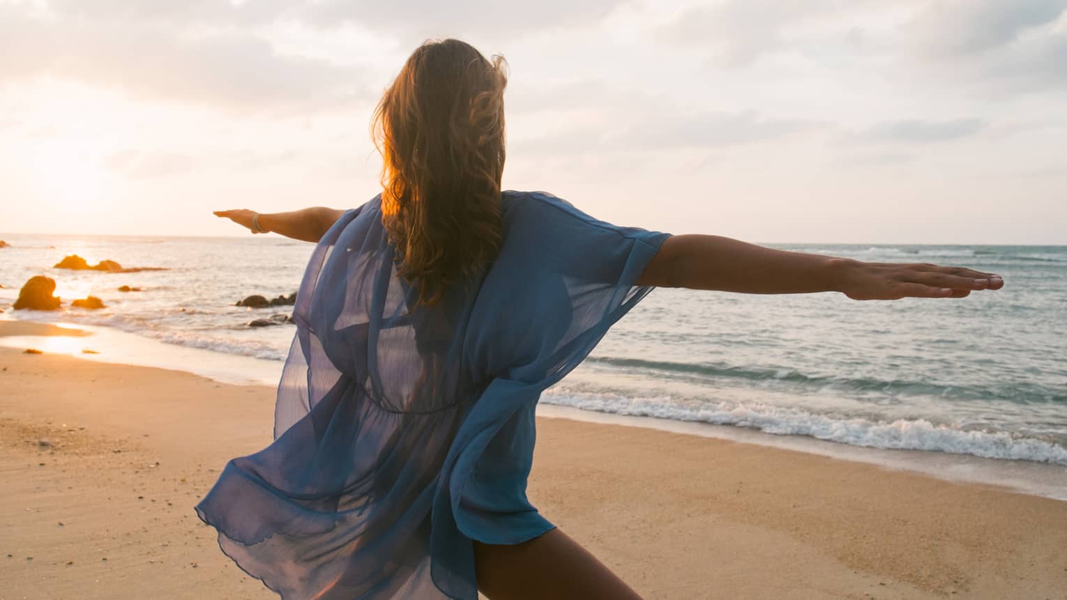 Woman wearing sheer beach cover kneels with arms outstretched in yoga pose on sandy beach by water