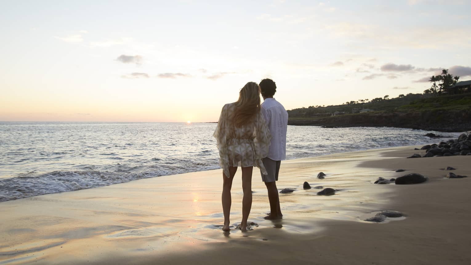 Back view of barefoot couple on sand beach leaning against each other, looking out at sunset over ocean