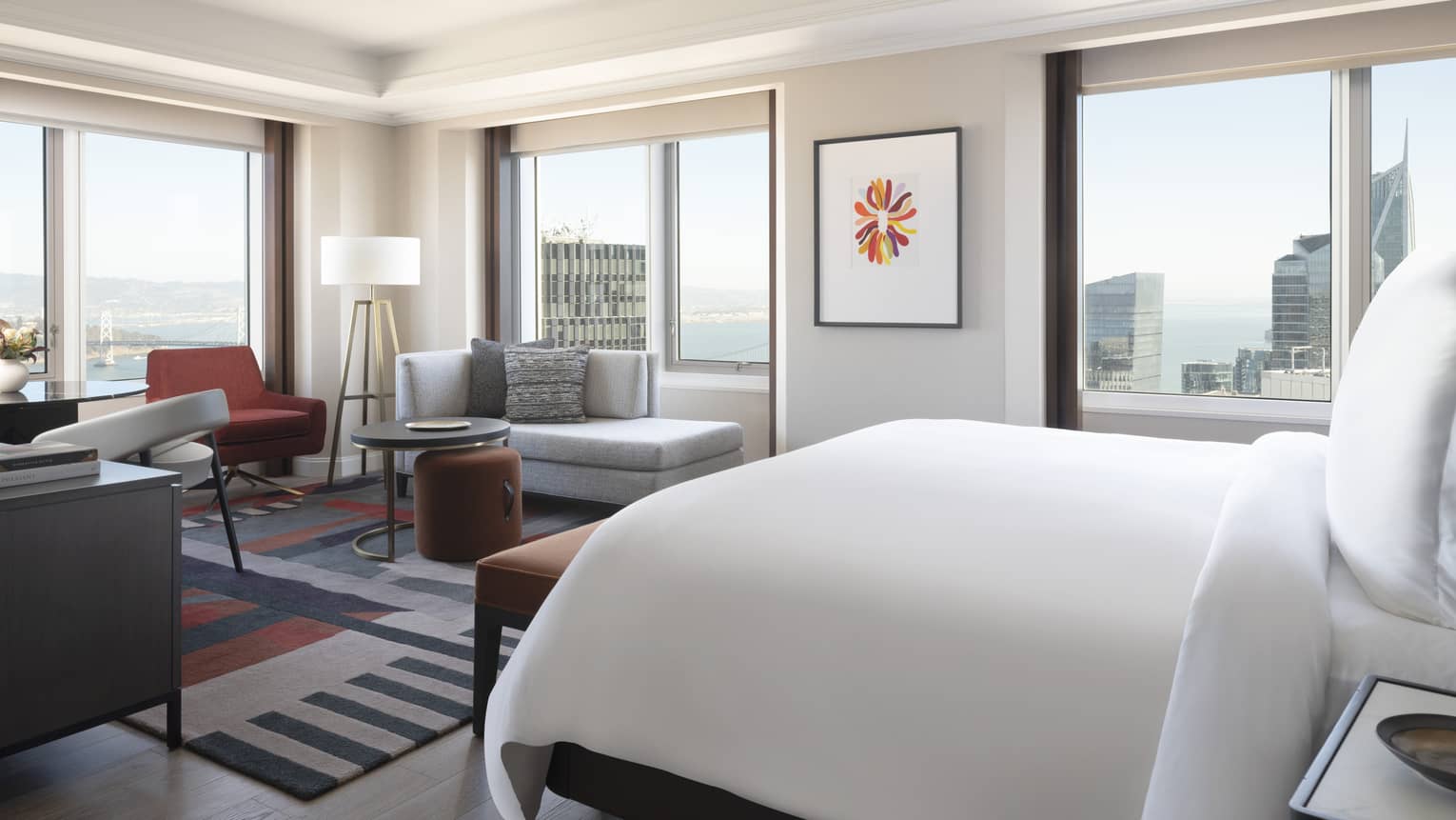 A hotel room with a large bed, large windows on each wall, a sofa, desk area, and lamp.