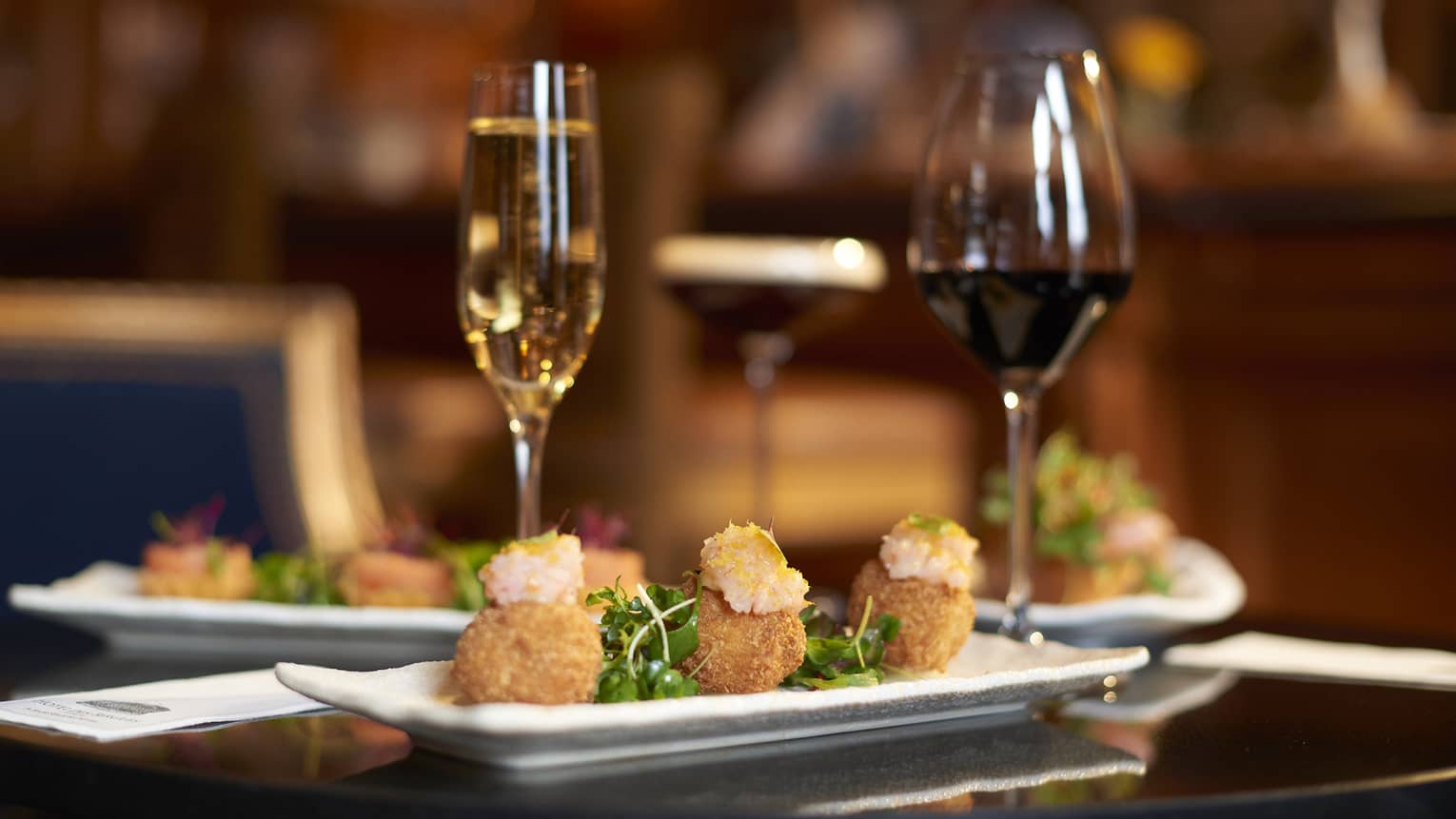 Three arancini on a white rectangular plate on a restaurant table with two glasses of wine