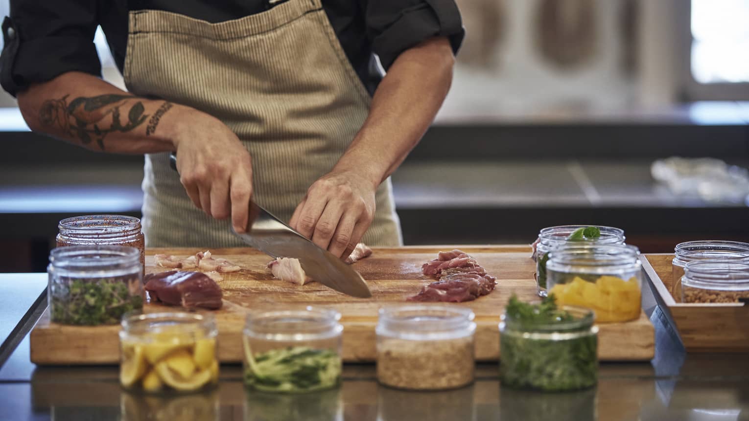 Close-up of chef slicing raw fish on wood butcher's block by glass jars with garnishes 