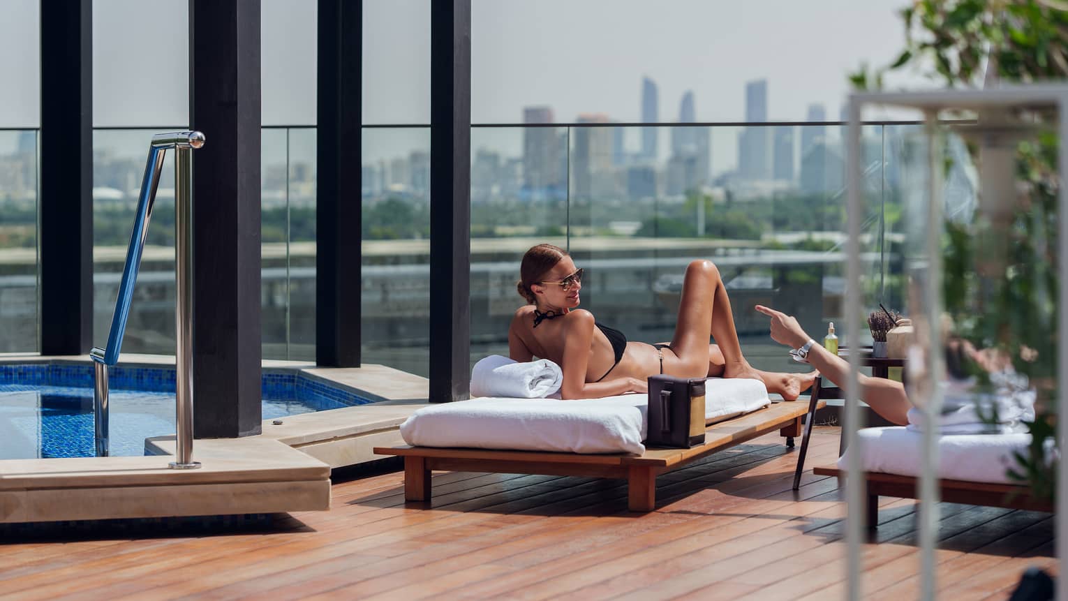 Person wearing black bikini lounges in the sun on a daybed next to a whirlpool on a rooftop deck with the city skyline in the background