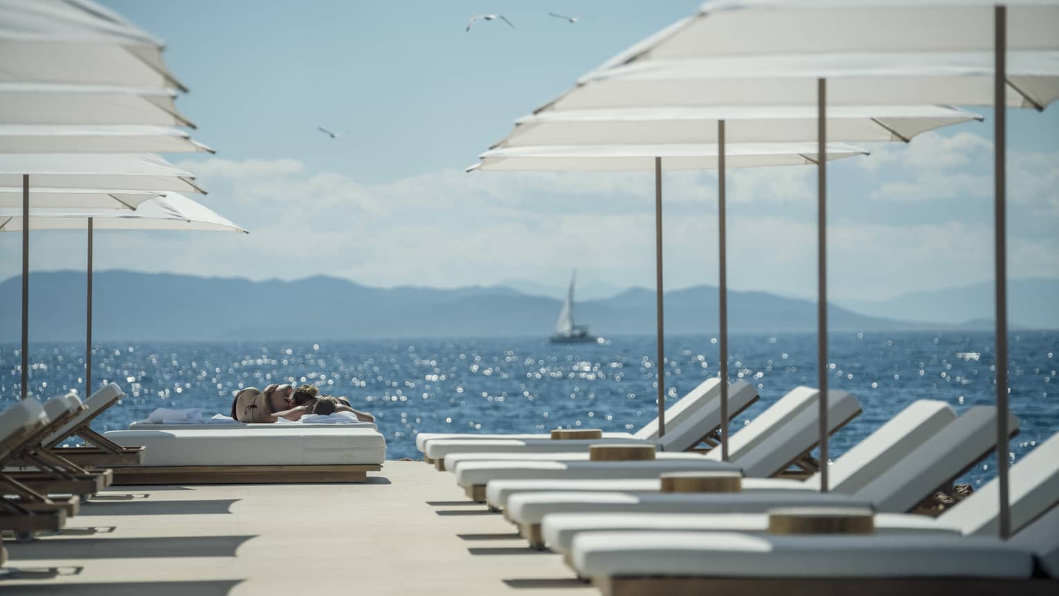 A sail boat sails in the distance while white lounge chairs are ready for guests and lined up next to the oceanside Nafsika Pool in Athens. 