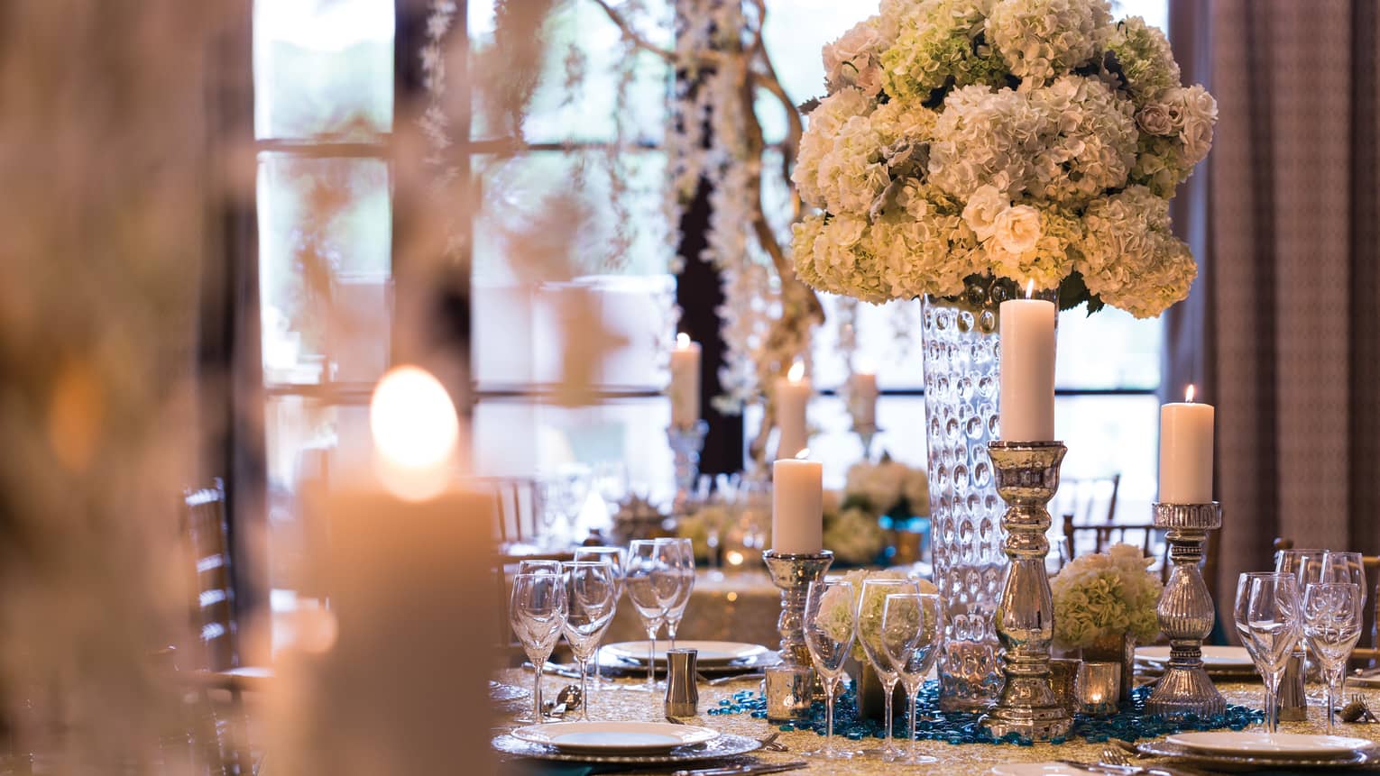 Close up of table set with glass vase, flowers, pewter candlesticks, candles, white plates