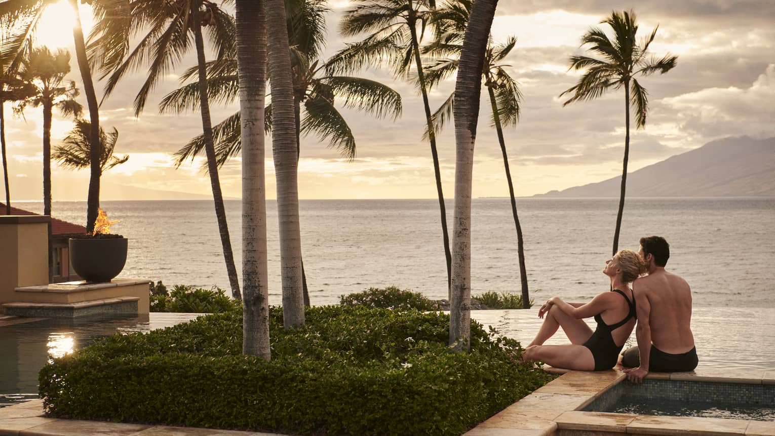Couple relaxing by an infinity pool with palm trees, overlooking the ocean at sunset with mountains in the background