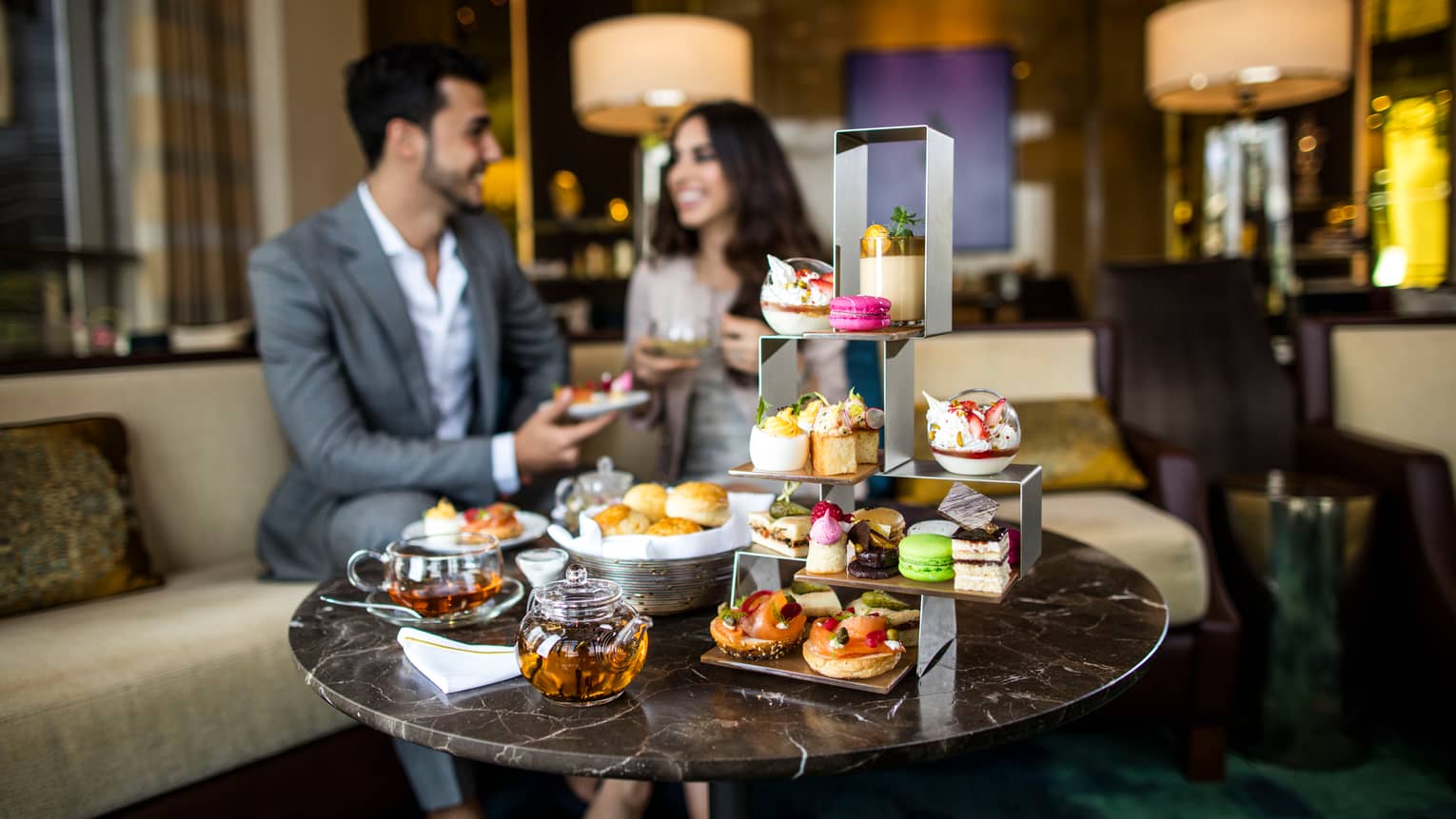 Coffee table with afternoon tea service, three-tiered tray with fine desserts, couple on sofa in background