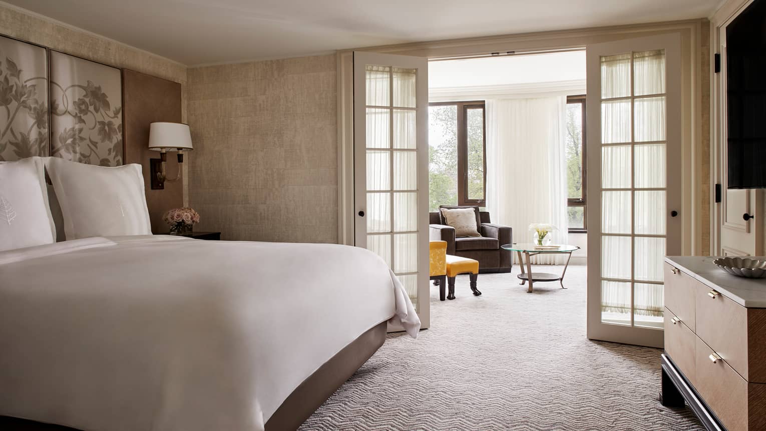 Senator Suite bed with white linens beside glass French doors leading to sunny living room with armchairs