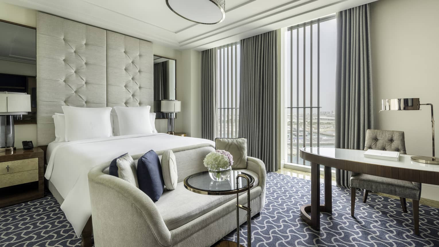 Hotel room with king bed, ceiling-height upholstered headboard, beige chaise, desk and floor-to-ceiling windows