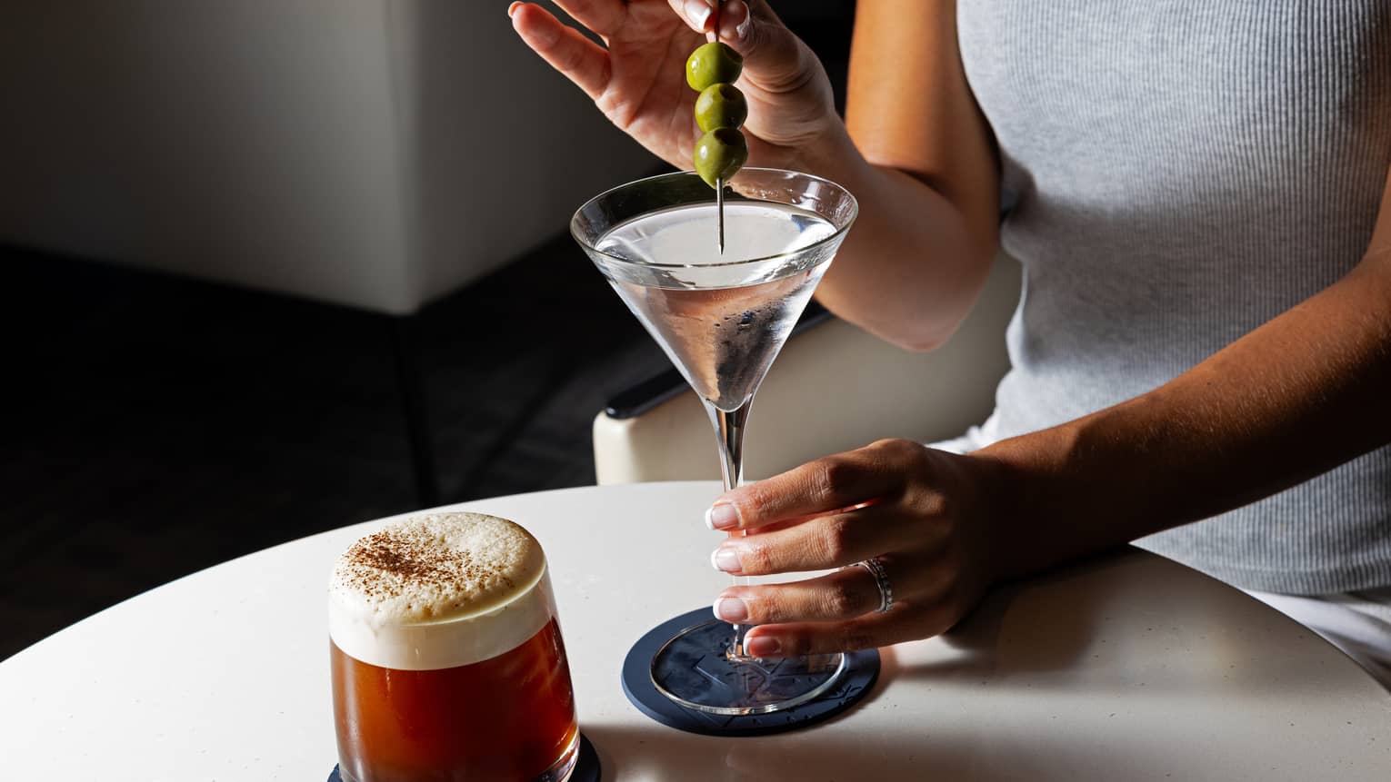 A person adds garnish of olives to a clear cocktail in a stemmed glass, next to an amber-coloured cocktail with a white foam top in a tumbler.
