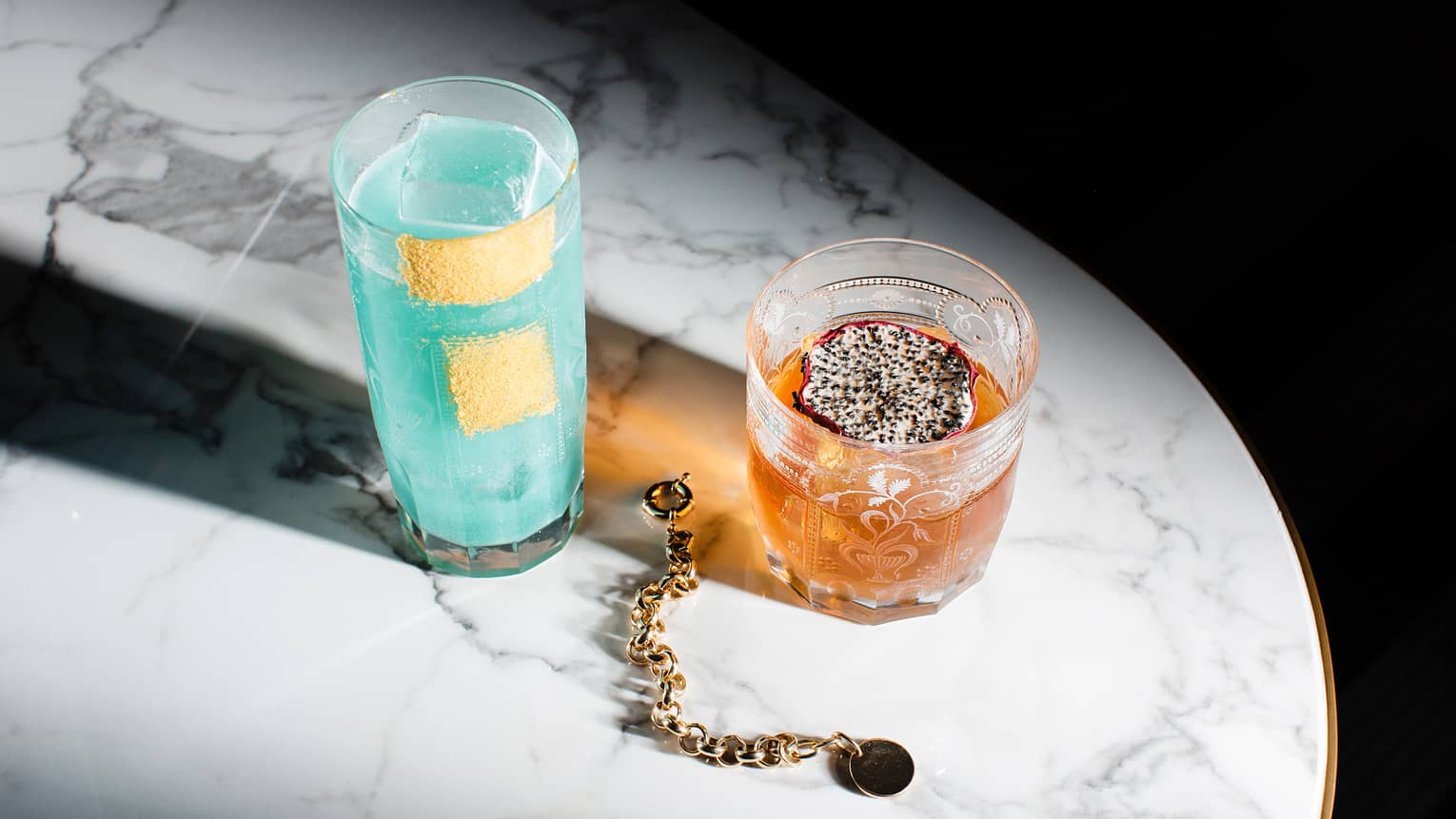 Light-blue cocktail in highball glass beside orange cocktail garnished with dragon fruit slice in old-fashioned glass on marble table