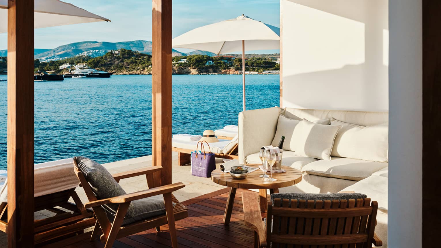 Cabana interior with white banquette-style couch, round teak table and two teak chairs open to platform with white umbrellas and blue sea views