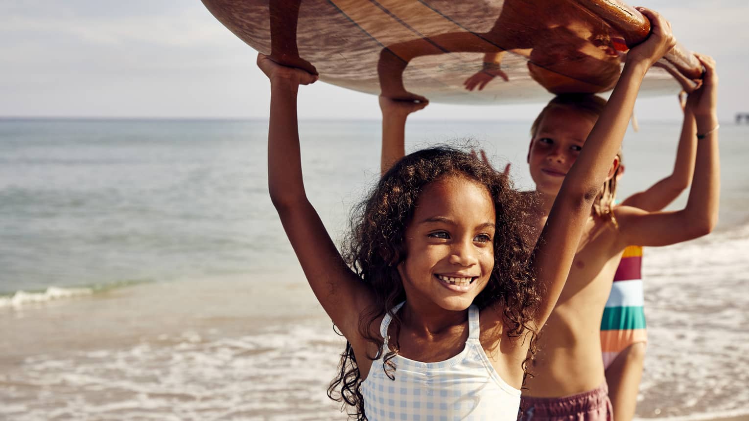 Three young children carry a surfboard on the beach, smiling and enjoying the sunny day. The ocean is in the background.