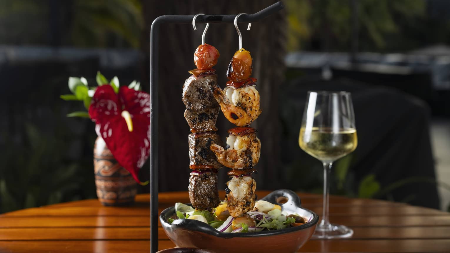 One shrimp skewer and one beef tenderloin skewer hang from a black serving bar next to a clay bowl filled with salad and a glass of white wine in the background