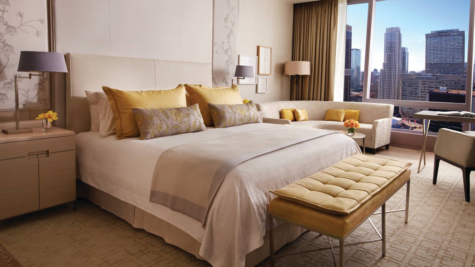 Plush yellow bench at foot of bed with yellow, gold pillows by curved loveseat, window