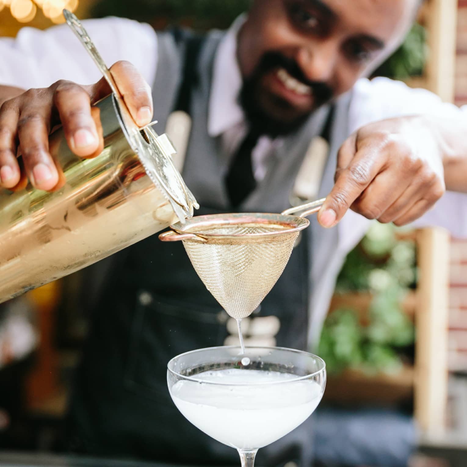 A smiling bartender pours a pale silvery liquid from a shaker through a strainer into a coupette martini glass.