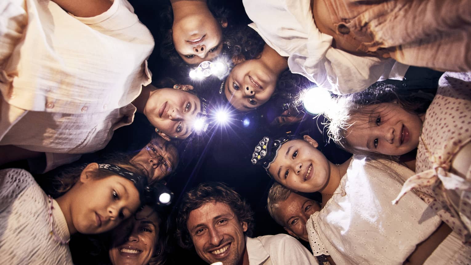 A group of children and adults, some wearing headlamps, stand in a circle and smile downwards into the camera. 