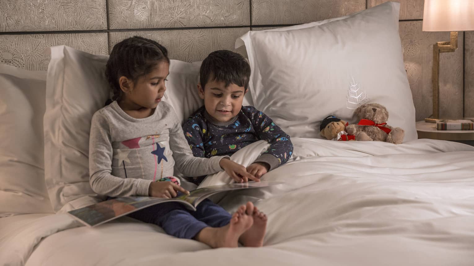 Young girl reads to her brother at bedtime, both in pajamas, a teddy bear propped on the pillow next to them