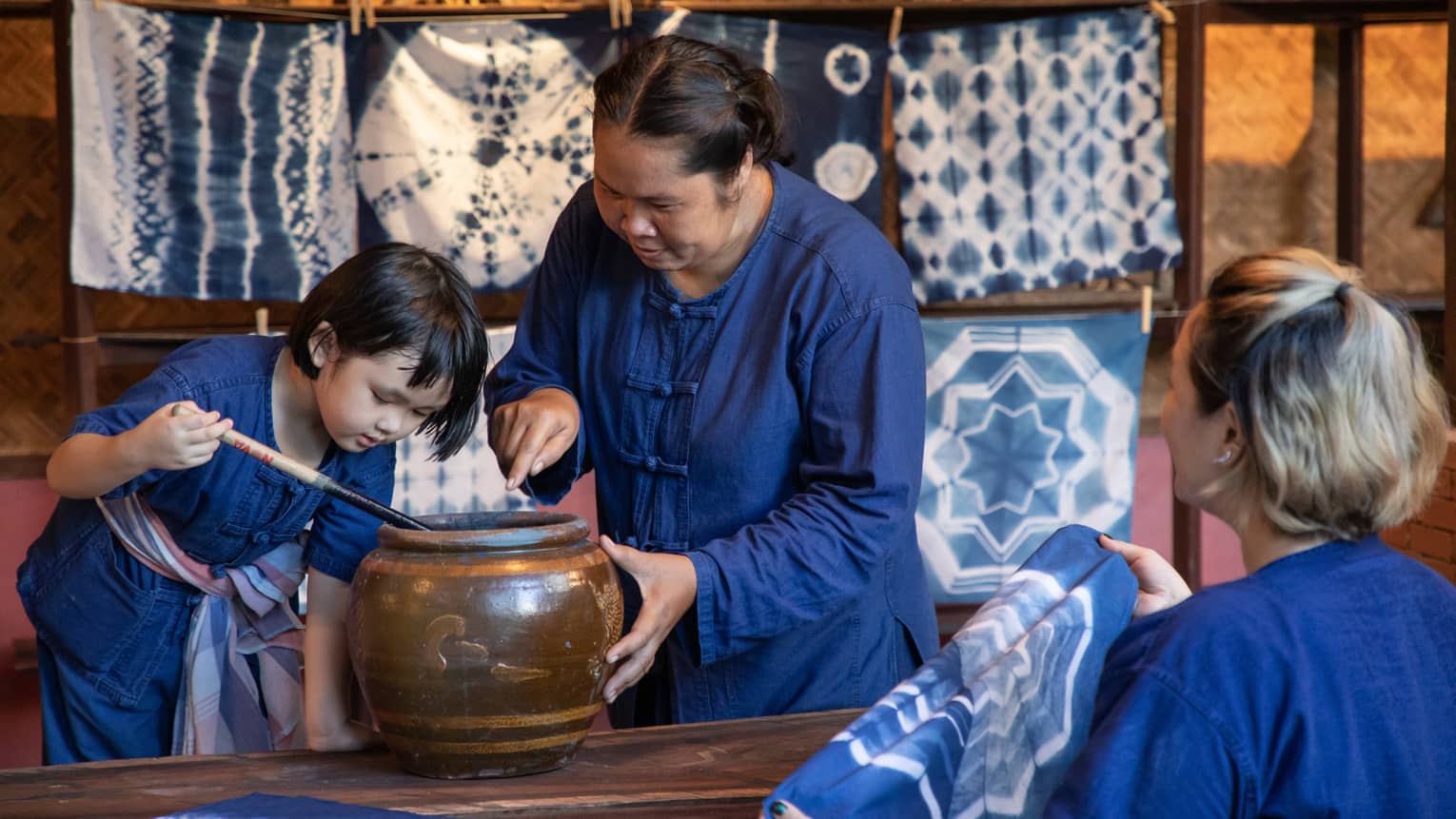 A woman teaching a child how to dye fabric using indigo, with tie-dyed cloths hanging in the background in a traditional workshop.