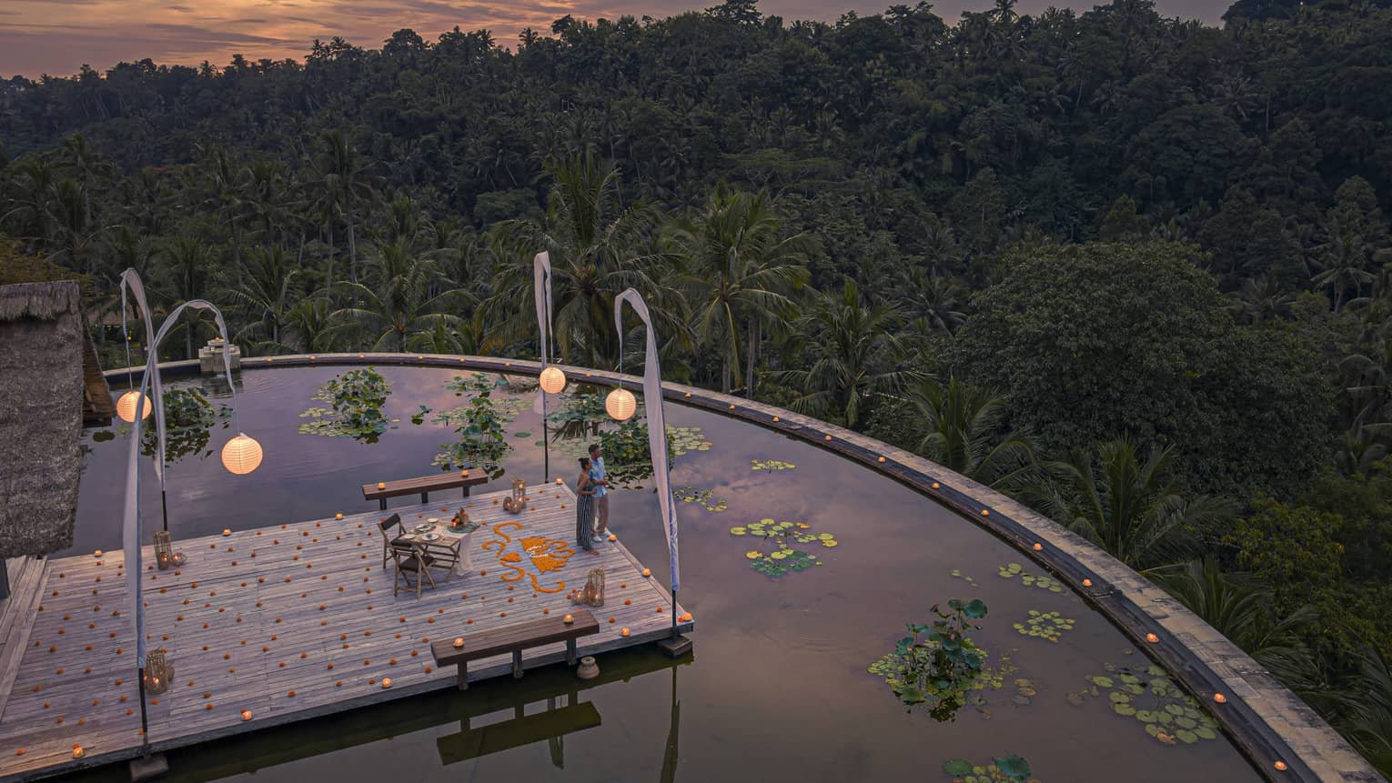 Couple stands on platform over lotus pond at dusk, dining table and hanging lanterns surrounding it, views of lush forest in front of them
