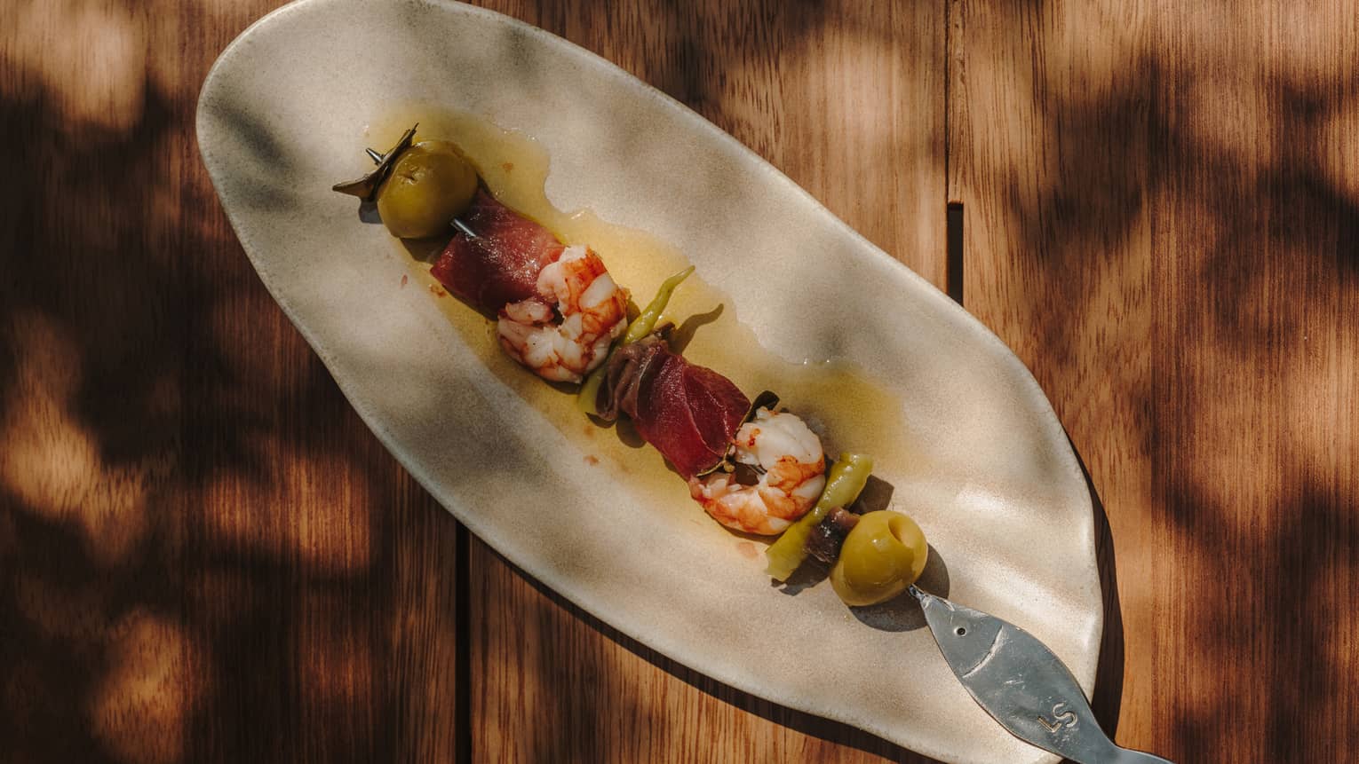 An artistic oval off-white plate holds a skewer of food items, including prawns and olives