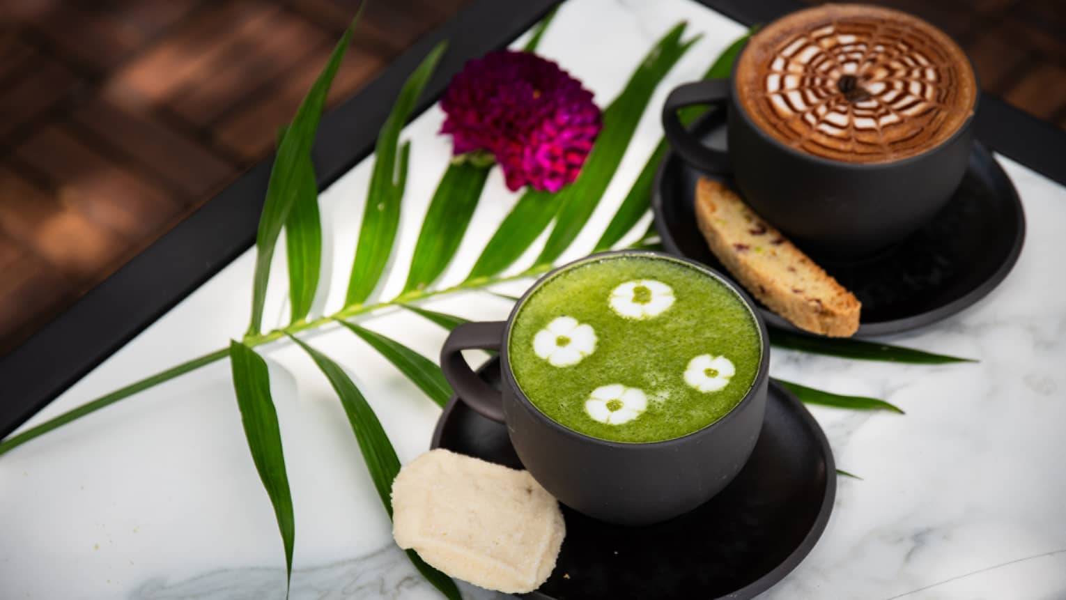 A black mug of coffee with a latte and brown-and-white foam and another black mug with tea with green foam and four white floral patterns